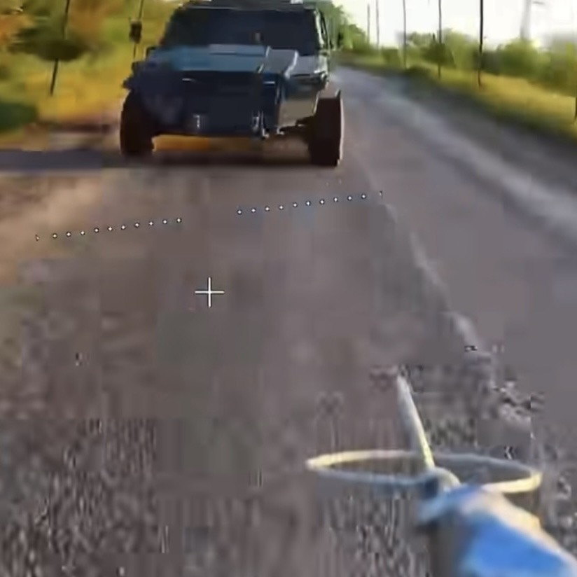 First-person view from an FPV drone camera on a low, fast approach along a dirt road toward an armoured vehicle.